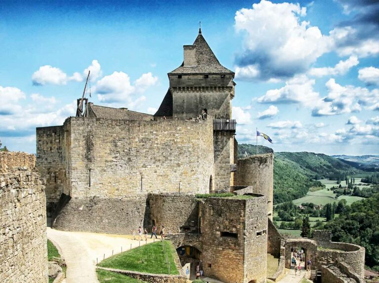 Panoramic view of Château de Castelnaud, a historic medieval castle in Dordogne, France, with tourists exploring the stone battlements under a blue sky