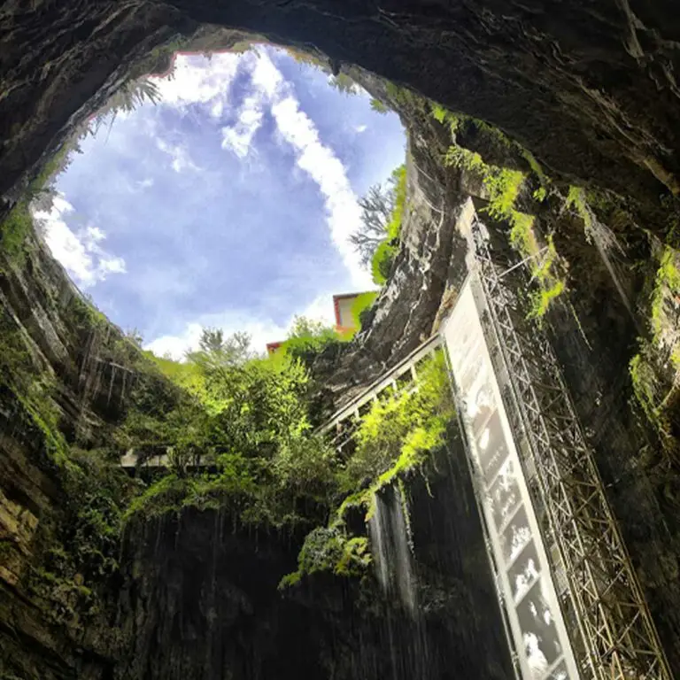 View from inside the Gouffre de Padirac cave looking up towards a blue sky through a natural circular opening, with cascading waterfalls and lush greenery on rocky walls, in France.