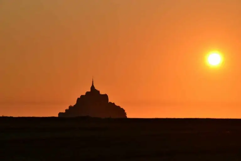 Silhouette of Mont-St-Michel at sunset with vibrant orange sky in Normandy, France, showcasing the iconic island commune during twilight.