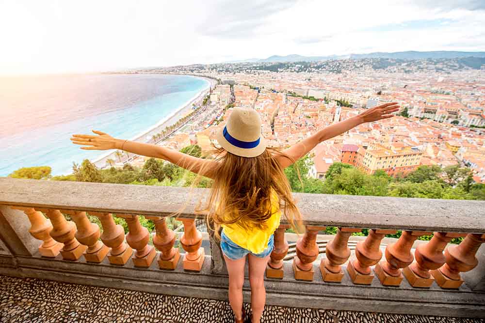young woman in yellow top standing with arms outstretched at a viewpoint in Nice, France