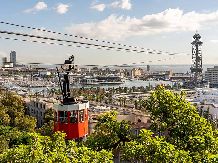 Red cable car ascending Montjuïc Hill with panoramic view of Barcelona port and cityscape, featuring lush greenery and historical tower.