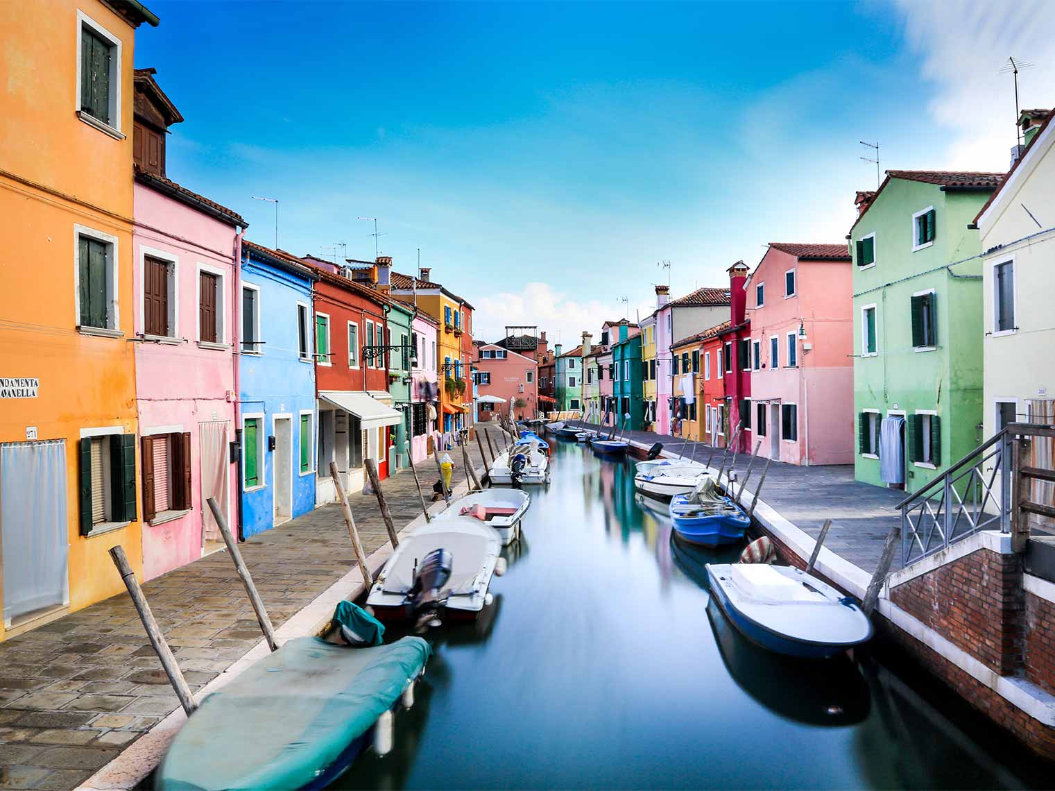 Colorful houses lining a serene canal with moored boats on Burano Island, Italy, showcasing traditional vibrant Venetian architecture and culture.