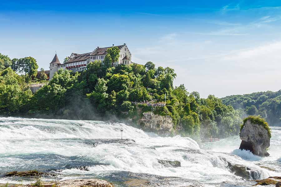 View of the Rhine Falls in Switzerland with a castle on the cliff and lush greenery under a clear sky