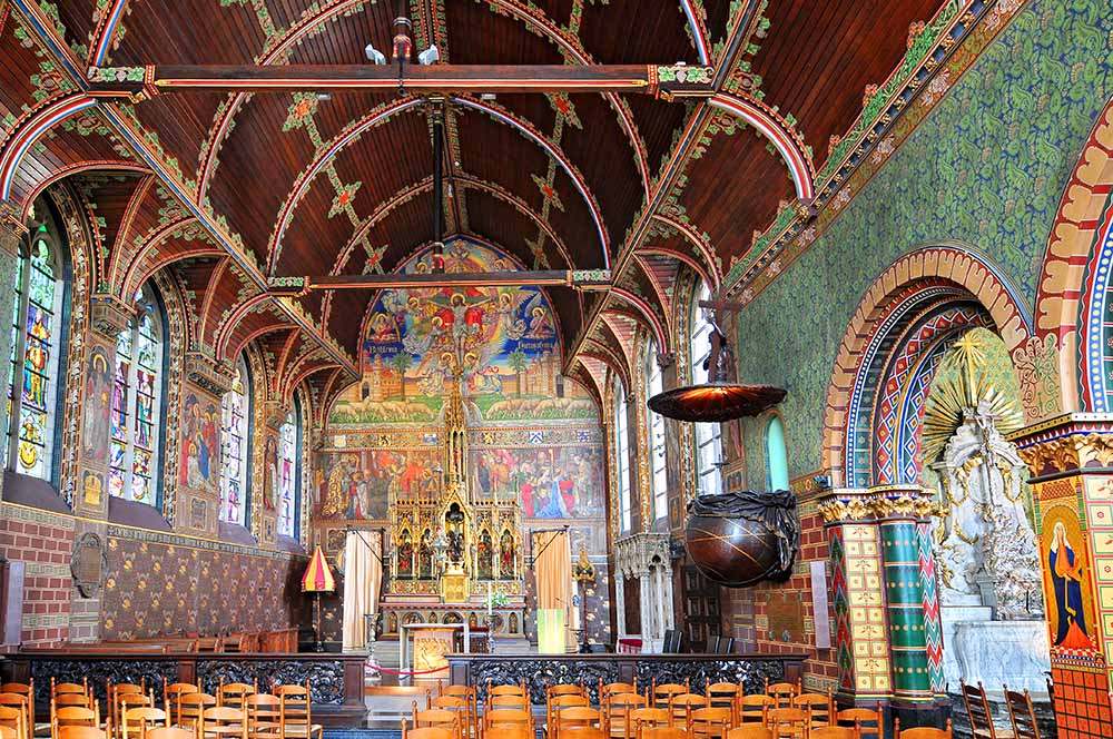 Interior of Basilica of the Holy Blood in Bruges, Belgium showing intricate stained glass, ornate altar, and colorful gothic revival architecture.